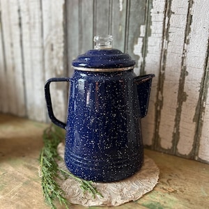 May include: A vintage navy blue enamelware coffee pot with white speckles. The pot has a glass lid and a black handle. It sits on a beige lace doily, with a sprig of greenery nearby. The background is weathered wood.