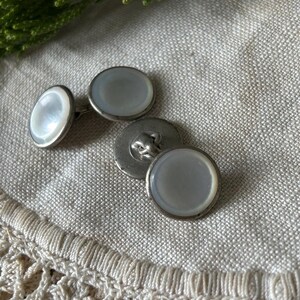 May include: A set of vintage cufflinks with a mother-of-pearl center and a silver-tone metal border. The cufflinks are oval and round, with a classic design. The background is a neutral linen fabric.