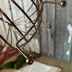 May include: A vintage, gold-toned metal wire basket with a spherical shape. The basket has a series of curved wires that intersect to form a cage-like structure. The base features three small, rounded feet. The overall design is retro and decorative.
