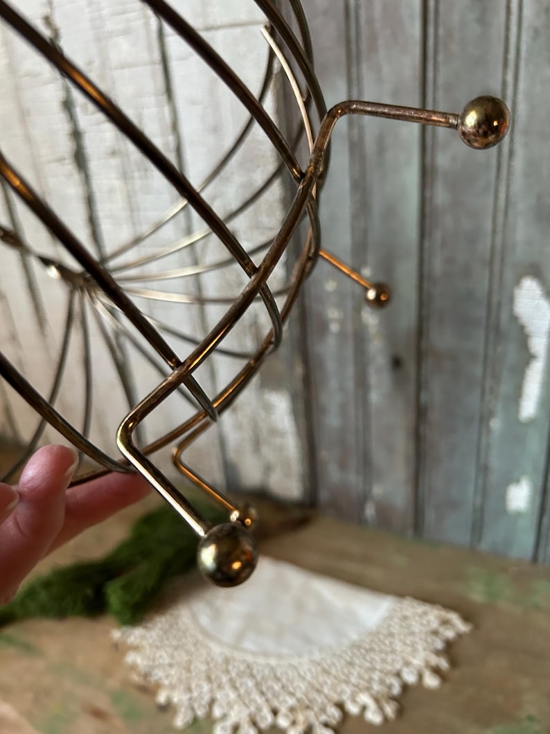 May include: A vintage, gold-toned metal wire basket with a spherical shape. The basket has a series of curved wires that intersect to form a cage-like structure. The base features three small, rounded feet. The overall design is retro and decorative.