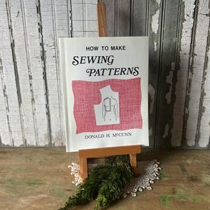 May include: A vintage book titled "How to Make Sewing Patterns" is displayed on a small wooden easel. The book cover features a red textured background with a diagram of a dress form. The author's name, Donald H. McCunn, is printed at the bottom.