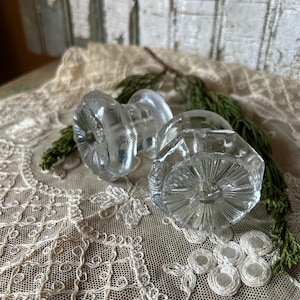 May include: Two clear glass knobs with a faceted design, resting on a cream-colored lace doily with floral patterns. The knobs are partially obscured by green sprigs, creating a vintage aesthetic.