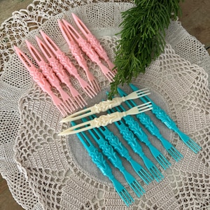 May include: A collection of decorative plastic appetizer forks in pink, white, and turquoise. The forks have ornate, vintage-style handles. The forks are arranged on a beige crocheted doily, with a sprig of green foliage.