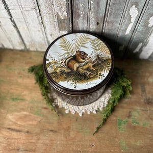 May include: Round metal tin with a detailed illustration of a chipmunk amidst ferns and foliage. The tin has a dark brown rim and a white background. The tin is placed on a white lace doily and green moss.
