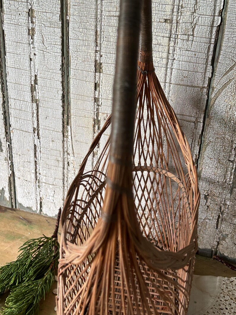May include: A close-up of a brown wicker basket with a tall handle. The basket has a woven diamond pattern and a rounded shape. A sprig of green foliage is visible on the left side of the image.