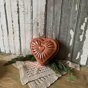 May include: A copper-colored heart-shaped mold with a decorative sunburst design. The mold sits on a beige crocheted doily with green sprigs. The background features weathered, distressed wooden planks.