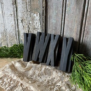 May include: A black wooden sculpture of the letters VWXY, resting on a cream-colored lace doily. The letters are thick and blocky, with a weathered appearance. Green sprigs of foliage are on either side of the sculpture, with a rustic wooden background.