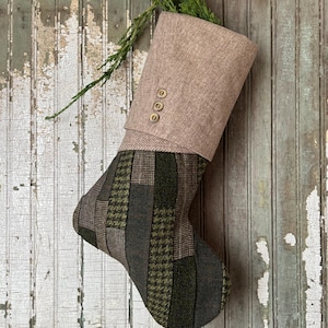 Pode incluir: Uma meia de Natal com um design de patchwork em tons de verde, castanho e cinza. A parte superior da meia é de tecido bege com três botões dourados. Ramos verdes são colocados dentro.