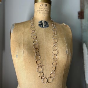 Puede incluir: Un collar largo de color plateado con una serie de círculos abiertos interconectados. El collar se exhibe en un maniquí de costura vintage con una cubierta de tela color beige. El maniquí tiene una cabeza de color latón y está sobre un fondo neutro.