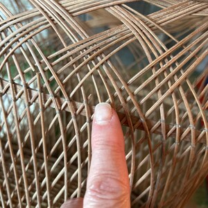 May include: Close-up of a woven wicker basket with a complex, crisscrossing pattern. The basket is made of light brown wicker and has a rounded shape. The texture of the wicker is visible, showing the natural material.