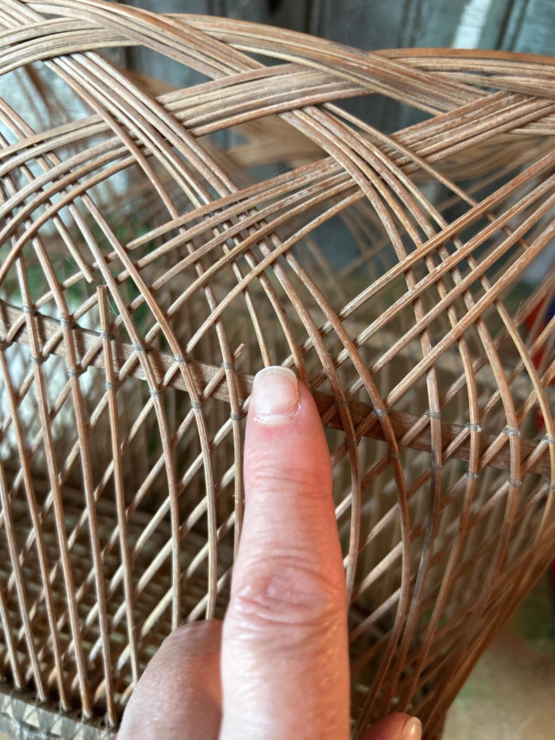 May include: Close-up of a woven wicker basket with a complex, crisscrossing pattern. The basket is made of light brown wicker and has a rounded shape. The texture of the wicker is visible, showing the natural material.