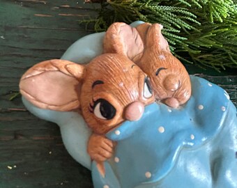May include: Two brown ceramic bunnies snuggled together under a blue blanket with white polka dots. The bunnies have closed eyes and are sleeping.