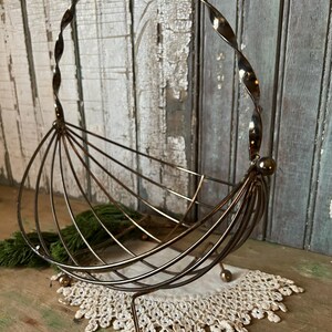 May include: A vintage metal wire fruit basket with a twisted handle. The basket has a rounded shape with vertical wire supports and rests on three small ball feet. It sits on a white lace doily.