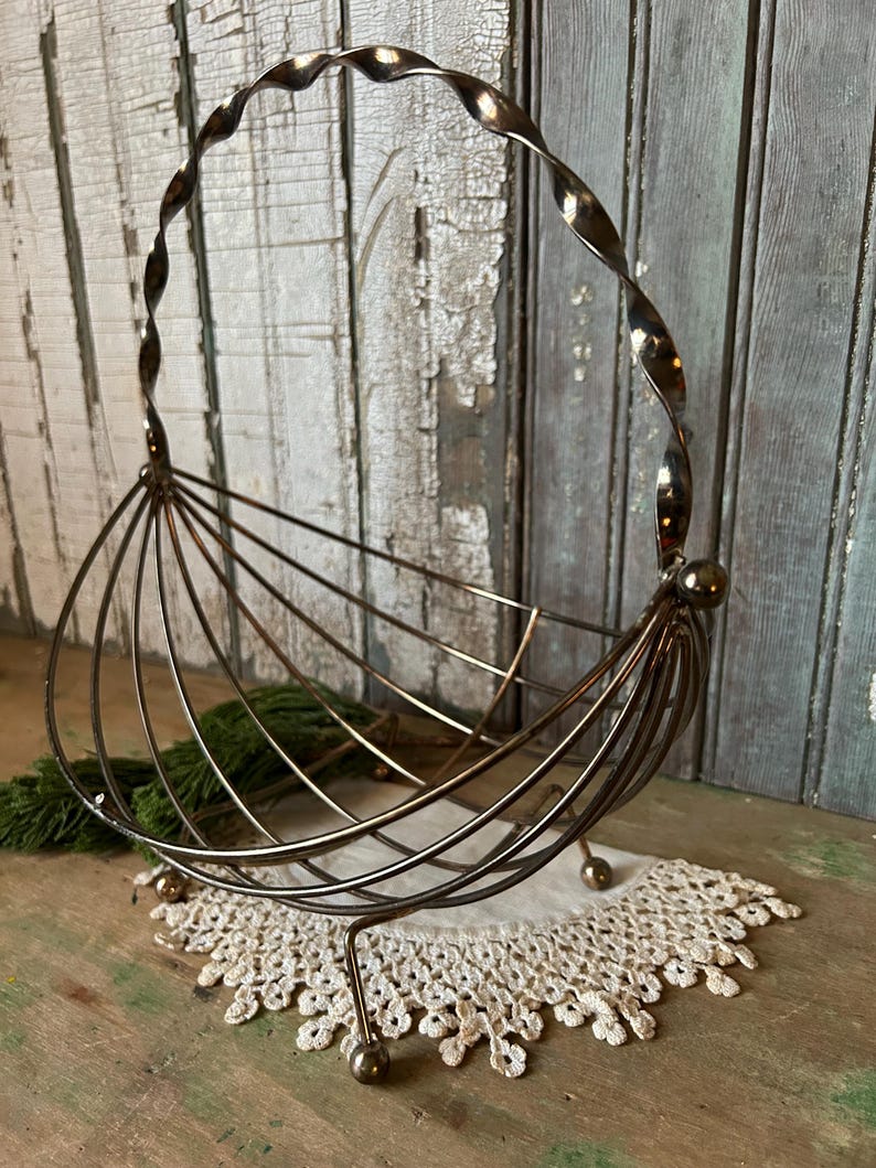 May include: A vintage metal wire fruit basket with a twisted handle. The basket has a rounded shape with vertical wire supports and rests on three small ball feet. It sits on a white lace doily.