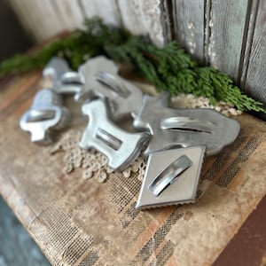 May include: A set of vintage metal cookie cutters in various shapes, including a star, a bell, a dog, and a rectangle. The cutters are silver in color and have a worn, antique look. They are arranged on a brown, textured surface.