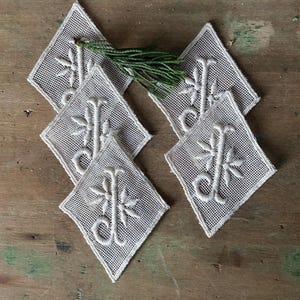 May include: Six diamond-shaped, off-white coasters with a floral and letter design. The coasters are made of a mesh material and are arranged on a wooden surface. A sprig of green foliage rests on top of the coasters.