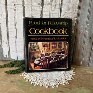May include: A vintage cookbook titled "Food for Fellowship Cookbook" by Antoinette Kuzmanich Hatfield. The cover is black with gold lettering and a picture of a dining table. The book is resting on a white lace doily.