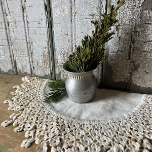 Peut inclure: Un petit vase en métal argenté avec un bord décoratif, contenant une branche de feuillage vert. Le vase est posé sur un napperon en lin blanc avec une bordure en dentelle crochetée. Le fond présente des planches de bois peintes en blanc et patinées.