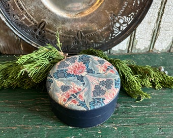 May include: A round, vintage trinket box with a floral pattern in shades of blue, pink, and green. The box is made of black cardboard and has a lid.