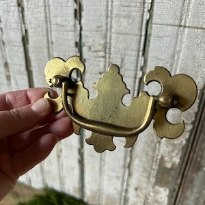 Könnte beinhalten: Antiker Schubladengriff aus Messing mit aufwendigem floralem Design. Der Griff hat einen gebogenen Griff und dekorative Endstücke. Die Hardware ist für einen Schrank oder eine Kommode geeignet.