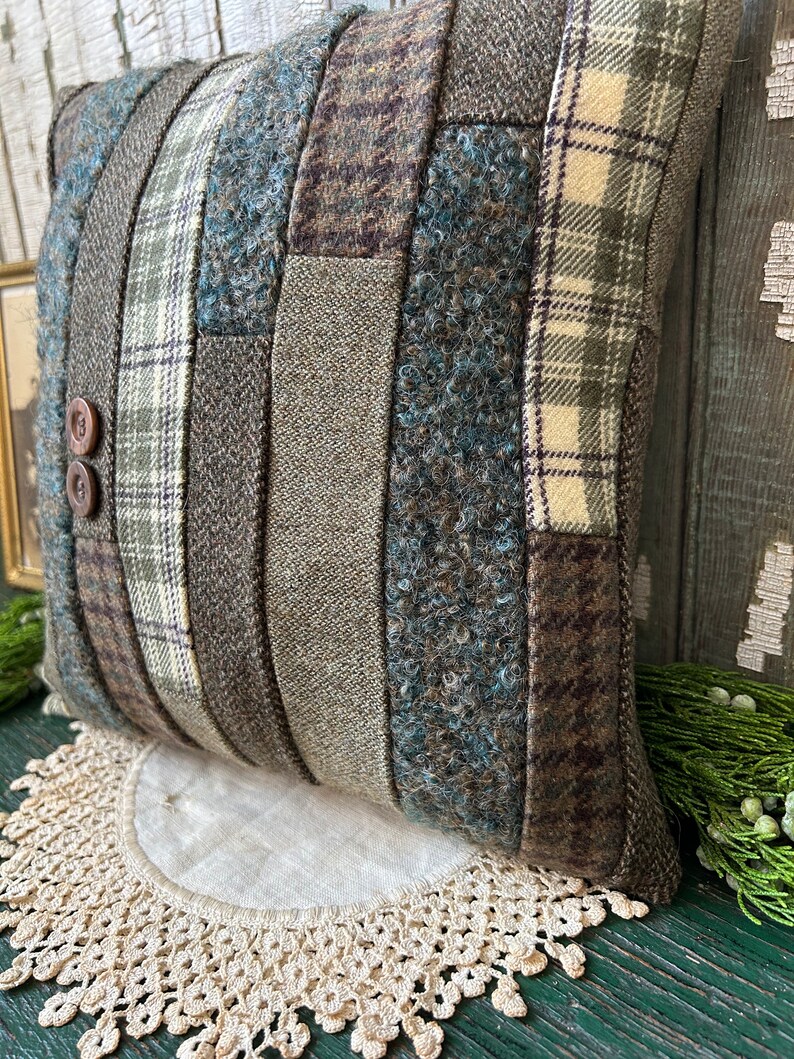 K&ouml;nnte beinhalten: Ein braunes und gr&uuml;nes Patchwork-Kissen mit Knopfdetail. Das Kissen liegt auf einer wei&szlig;en H&auml;kelspitze.