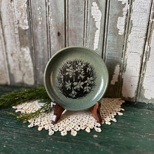 Pode incluir: Um pequeno prato de cerâmica verde com uma silhueta preta de um desenho floral. O prato está sobre uma renda branca e um suporte de madeira.