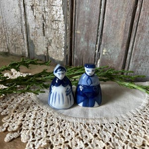 Könnte beinhalten: Zwei kleine Porzellanfiguren, die einen Mann und eine Frau darstellen. Die Figuren sind hauptsächlich blau und weiß, mit detaillierter Kleidung und Gesichtszügen. Sie stehen auf einer Spitzendecke vor einem rustikalen Holzhintergrund und erzeugen ein Gefühl von Nostalgie.