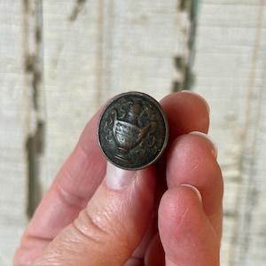 May include: A small, antique bronze button with an embossed design. The button features a central urn motif with decorative elements. The button is round and has a slightly tarnished appearance, suggesting age. The button is held between fingers.
