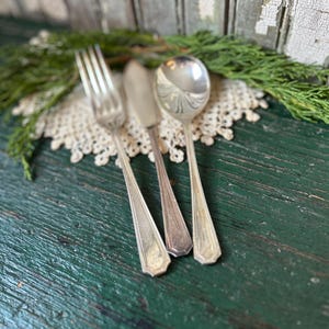 May include: Three silver-plated utensils, a fork, a butter knife, and a spoon, are arranged on a white crocheted doily. The utensils are on a green wooden surface.