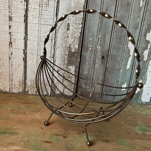 May include: A vintage metal wire basket with a twisted handle and a curved, open design. The basket is supported by three small ball feet and is made of a bronze-colored metal. The background features weathered wooden planks.