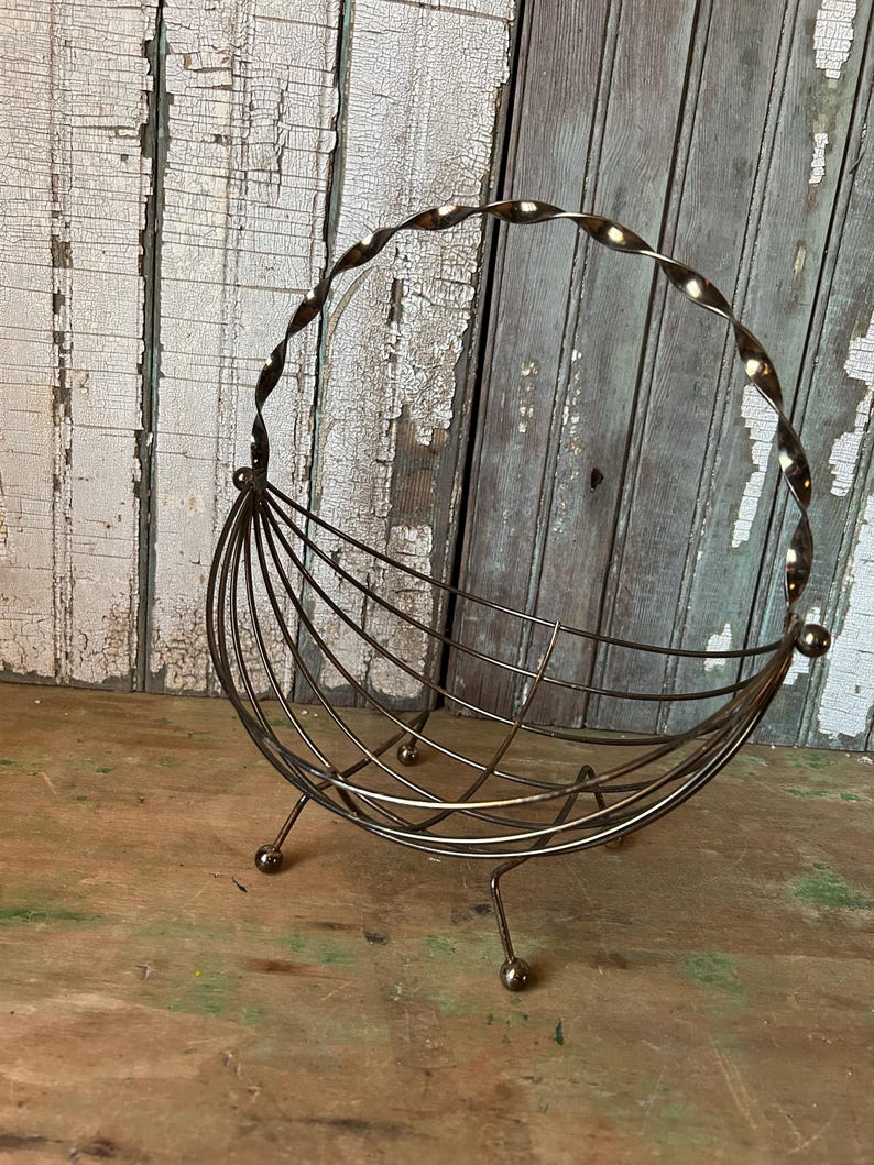 May include: A vintage metal wire basket with a twisted handle and a curved, open design. The basket is supported by three small ball feet and is made of a bronze-colored metal. The background features weathered wooden planks.