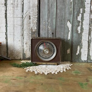 May include: A vintage brown and gold Taylor barometer with a circular gauge and a needle indicating the current atmospheric pressure. The barometer is encased in a rectangular frame and sits on a small stand. A sprig of greenery and a lace doily are in the foreground.