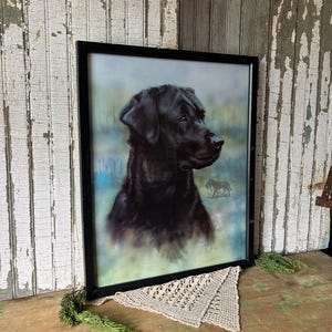 Pode incluir: Retrato emoldurado de um cão Labrador Retriever preto. A obra apresenta uma representação detalhada da cabeça e ombros do cão contra um fundo suave e desfocado. A moldura preta adiciona elegância clássica.