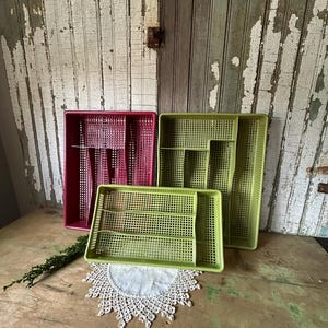 May include: Three plastic utensil organizers in shades of pink and green. The organizers have a grid-like design and are of varying sizes. They are arranged on a wooden surface with a lace doily and greenery.