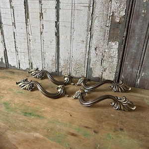 May include: A set of four antique brass-toned metal drawer pulls featuring an ornate, curved design with detailed leaf-like flourishes at each end. These hardware pieces have a smooth, metallic finish and a distinct arch shape for mounting.