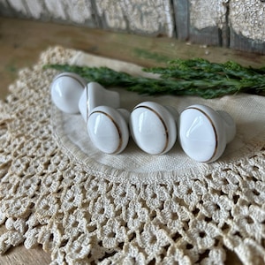 Könnte beinhalten: Fünf weiße Keramik-Schrankknöpfe mit goldfarbenem Rand, angeordnet auf einer cremefarbenen Häkelspitze. Der Hintergrund zeigt eine Holzoberfläche und grüne Zweige, was eine Vintage-Ästhetik erzeugt. Jeder Knopf hat einen goldenen Streifen.