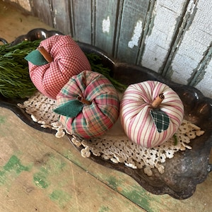 May include: Three fabric apples in red and green patterns rest on a silver tray with a lace doily. The apples have cinnamon stick stems and green fabric leaves. The background features weathered, painted wood.