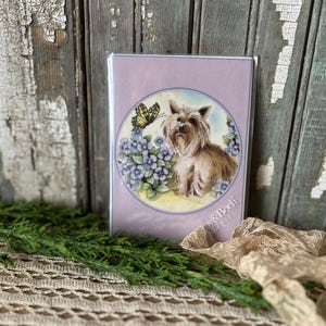 May include: A greeting card with a Yorkshire Terrier illustration. The artwork shows the dog with blue flowers and a yellow butterfly on a lavender background. The card is displayed on a rustic wooden surface.
