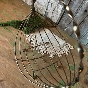 May include: A vintage-style metal wire basket with a twisted handle. The basket has a cage-like design with a rounded bottom and small ball feet. A white lace doily and a sprig of greenery are inside.