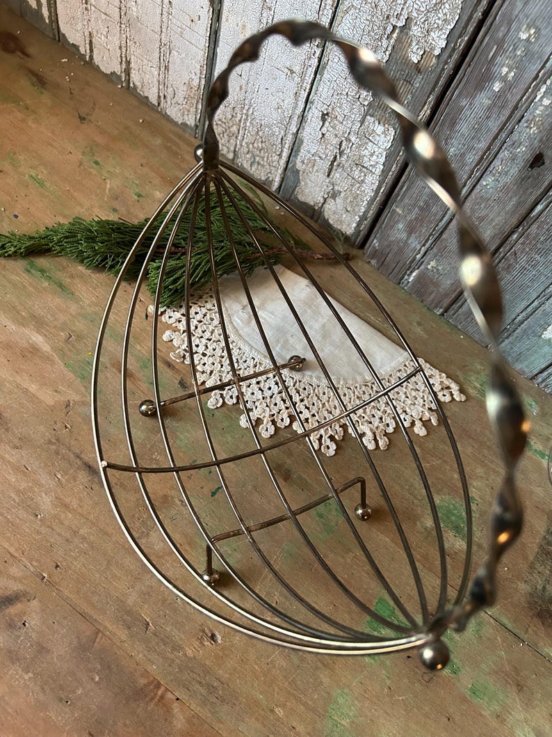 May include: A vintage-style metal wire basket with a twisted handle. The basket has a cage-like design with a rounded bottom and small ball feet. A white lace doily and a sprig of greenery are inside.