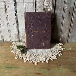 May include: A vintage book with a dark purple cover and gold lettering, titled "Spiegel der Taufe." The book rests on a cream-colored, crocheted doily with a sprig of greenery. The background features weathered, white-painted wood.