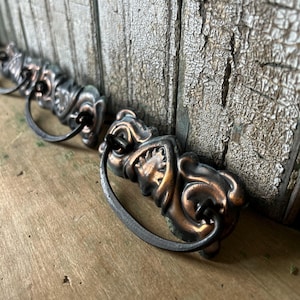 May include: A set of two metal drawer pulls featuring a dark copper finish with ornate, raised leaf-like detailing and a curved, dark-toned drop handle attached to each rectangular base plate, showcasing a weathered and textured appearance.