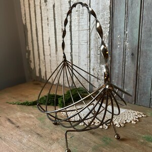 May include: Vintage metal basket with a twisted handle and a fan-like design. The basket has a bronze finish and is supported by three small legs with ball feet. The handle is a decorative element.
