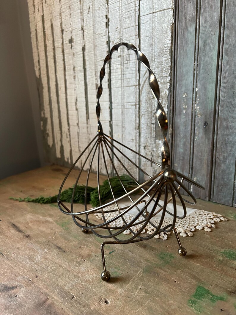 May include: Vintage metal basket with a twisted handle and a fan-like design. The basket has a bronze finish and is supported by three small legs with ball feet. The handle is a decorative element.