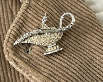 May include: A silver-toned brooch in the shape of a genie lamp, encrusted with clear rhinestones. The lamp is set against a brown corduroy fabric, showcasing its intricate design and sparkling details. The brooch has a curved handle and a flame.