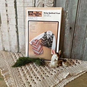 May include: A pattern for a strip quilted vest, with a photo of the finished product. The pattern is titled "Strip Quilted Vest" and includes sizing information. A small ceramic deer figurine and a sprig of greenery sit on a crocheted lace doily.