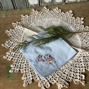 Può includere: Un fazzoletto azzurro con cani dei cartoni animati ricamati, appoggiato su un tovagliolo di lino beige e un centrino di pizzo color crema. Un rametto di fogliame verde è incluso. Su una superficie di legno.