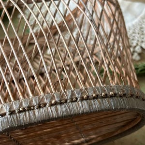 May include: A close-up of a woven, dome-shaped basket. The basket is made of light brown, thin wooden strips arranged in a lattice pattern. The base is reinforced with a darker, woven material and small wooden beads.