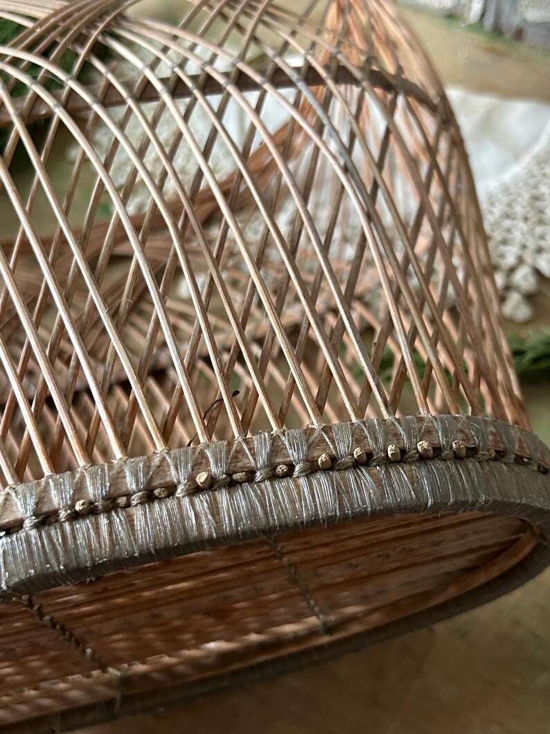 May include: A close-up of a woven, dome-shaped basket. The basket is made of light brown, thin wooden strips arranged in a lattice pattern. The base is reinforced with a darker, woven material and small wooden beads.