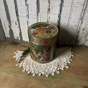 May include: A vintage round tin with a green and gold design. The tin features a detailed illustration of a European town on the lid and figures in period clothing around the sides. It sits on a white lace doily.